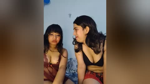 Video of two South Asian women in lingerie, sitting on a bed with patterned sheets. Both wear black bras; one has long dark hair and the other has short dark hair.
