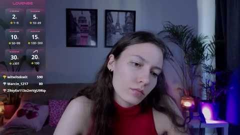 Video of a young woman with long brown hair, wearing a red top, sitting on a bed with a purple pillow. Background includes a plant, framed art, and a digital screen displaying social media stats.