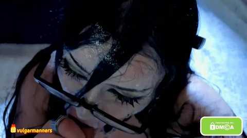 A close-up video of a young woman with long, wet black hair, wearing glasses, lying in a bathtub, her eyes closed. The image has a dim, blue-tinted lighting.