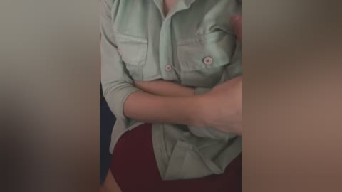 Video of a person wearing a light green, button-down shirt and burgundy shorts, arms crossed, with a blurred background.