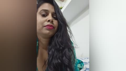Video of a South Asian woman with medium-dark skin, long black hair, and a teal floral blouse. She has a neutral expression and is indoors, possibly in a bedroom.