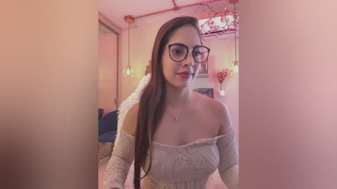 Video of a young woman with long brown hair, wearing glasses and a cream off-shoulder sweater, smiling in a pink-themed room with heart-shaped lights.