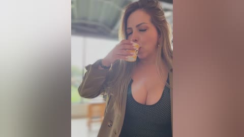 Video of a curvaceous, fair-skinned woman with long, wavy blonde hair, wearing a low-cut, dark green patterned top, eating a sandwich. She's in a bright, modern indoor setting with large windows.