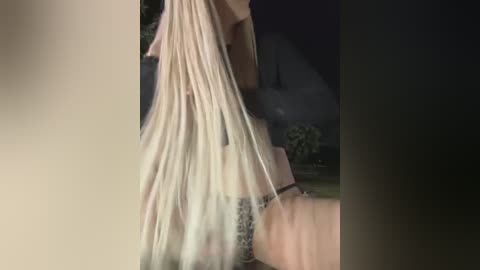 Video of a blonde woman with long, straight hair, wearing a black lace bra, standing outdoors at night. The background is blurred with a dark, indistinct house and greenery.