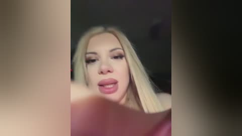 A blurry, low-quality video of a young woman with long, straight blonde hair, wearing heavy makeup and a pink top, with a dimly lit, indistinct background.