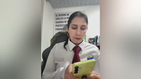 Video of a Latina woman with dark hair in a bun, wearing a white shirt, red tie, and hoop earrings, reading a green phone in a small, white-walled office.