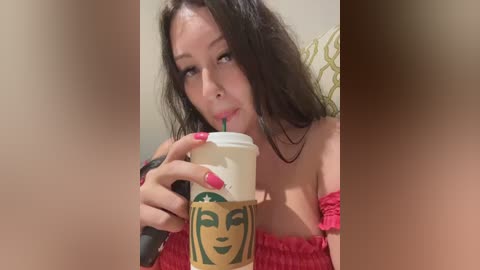Video of a young woman with long black hair, wearing a red off-shoulder top, drinking from a Starbucks cup with a green logo, indoors with blurred background.