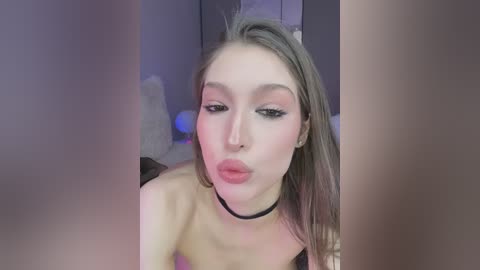 Media: Video of a fair-skinned, blonde-haired woman with bold makeup, pouting, wearing a black choker. The background is blurred, suggesting an indoor setting.