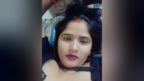 A video of a woman with medium-dark skin and black hair, wearing headphones, sitting with a contemplative expression, a nose ring, and a red dot on her forehead, blurred background.