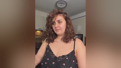 Video of a middle-aged woman with curly brown hair, wearing a black spaghetti-strap camisole with pink floral patterns, standing in a modern kitchen with white cabinets and stainless steel appliances.