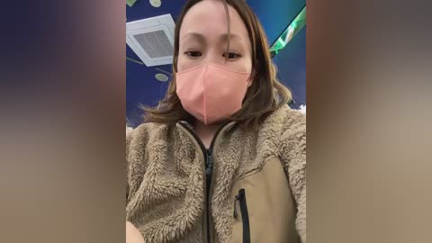 Video of a woman with light skin and brown hair, wearing a beige fleece jacket, pink face mask, and a white surgical mask. The background features a blue ceiling with circular lights and a green plant.