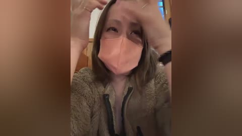 Video of a woman with light skin, wearing a pink face mask, brown hair, and a gray fur jacket, holding a glass in front of her face.