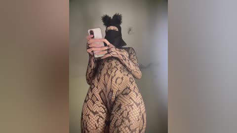 A video of a person in a black lace bodysuit with intricate patterns, black face mask, and black furry headband, holding a smartphone. The background is blurred and neutral-colored.