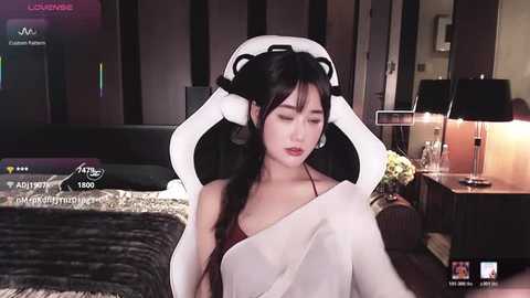 Video of a young Asian woman with long black hair, wearing a white blanket, sitting in a gaming chair in a dimly lit bedroom.