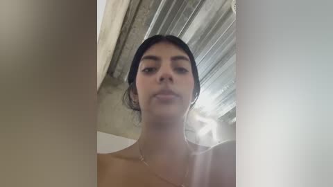 A close-up video of a young woman with medium skin tone, dark hair tied back, and a simple gold necklace. She gazes directly at the camera, her expression neutral. The background features a textured ceiling and a window with semi-transparent curtains.