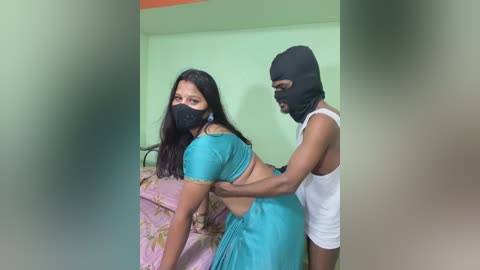 Video: A dark-skinned woman in a turquoise sari is restrained by a man in a black mask and white tank top. They stand in a green room with a floral-patterned bed in the background.