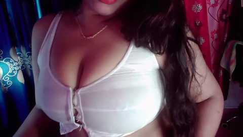 Video of a curvaceous woman with long, dark hair, wearing a white, lacy bra that accentuates her ample breasts, against a backdrop of colorful, patterned curtains.