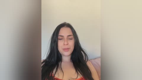 Video of a fair-skinned woman with long black hair, wearing a red lace bra, seated against a plain white wall, eyes closed in a serene expression.