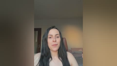 Video of a young woman with long, straight black hair, fair skin, and a slender build, sitting in a dimly lit room. She wears a white tank top, with a beige wall and a wooden door visible in the background.