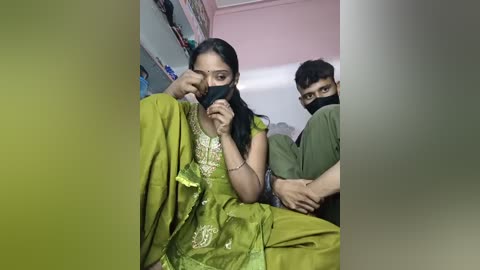 Video of a woman in a green sari with intricate embroidery, covering her face with a black mask, seated beside a man wearing a green mask, in a dimly lit room with shelves and pink walls.