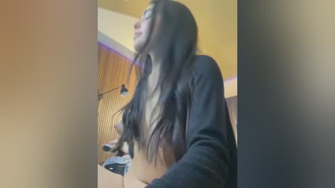 Video of a slender, topless woman with long dark hair, wearing a black cardigan, standing in a modern bathroom with wooden panels, yellow walls, and a ceiling light.