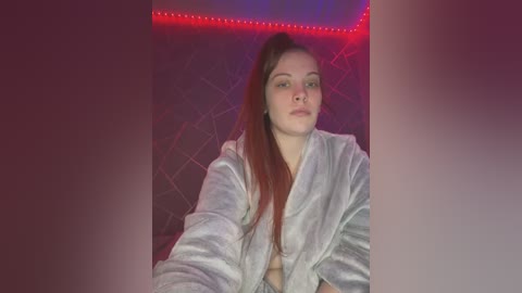 A video of a young, light-skinned woman with long red hair, wearing a fluffy white bathrobe, sitting against a dark quilted wall with red LED strip lighting.