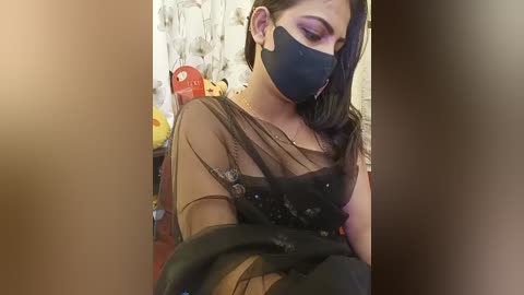 A video of a woman with medium skin tone and long, dark hair, wearing a black face mask, black top, and sheer black scarf. Background includes a yellow toy and floral wallpaper.