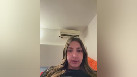 Video of a young woman with long, straight, blonde hair, wearing a black shirt, standing in a beige-walled room with an air conditioning unit above her.