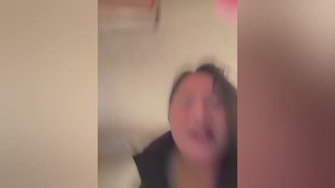 A blurry video of a woman with shoulder-length black hair and glasses, wearing a dark top, standing in a dimly lit room with beige walls and indistinct objects in the background.