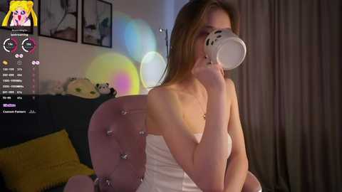 Video of a young, fair-skinned woman with long brown hair, wearing a white top, drinking from a white cup, seated on a pink, tufted chair in a dimly lit room with soft, pastel-colored lights and plush toys.