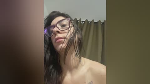 A video shows a wet, black-haired woman with glasses, wearing a black top, in a bathroom with a brown shower curtain.