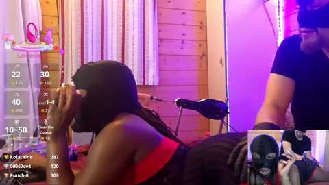 Video of a dark-skinned woman with long hair in a red bra, lying face down on a table in a cozy room with wooden walls, and a man in a black shirt standing nearby.