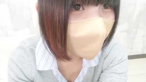 Video of an East Asian woman with shoulder-length, straight, dark hair, wearing a light gray sweater over a white collared shirt, and a beige surgical mask covering her nose and mouth. She has large, expressive eyes and a pale complexion. The background is a blurred, white, possibly hospital-like setting.