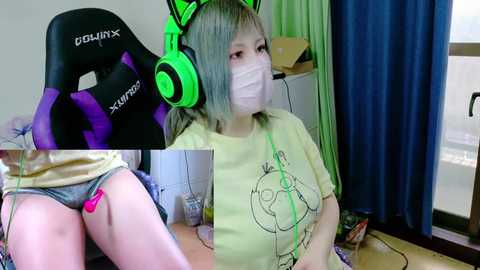 Video of an Asian woman with light skin, wearing green headphones, a yellow T-shirt, and a face mask, sitting on a desk chair. Background shows a gaming setup with a blue curtain and a \"CDLUX\" gaming chair.