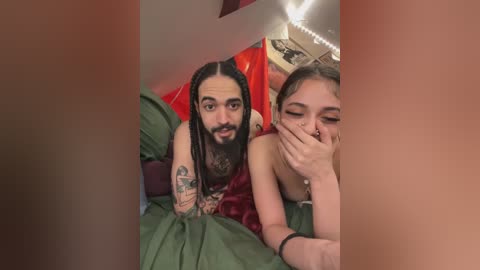 Video of a tattooed man with long hair and a woman with a nose ring, both on a green bed, laughing, covered by a red blanket.