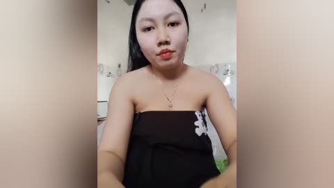 A video of an Asian woman with fair skin and long black hair, wearing a strapless black dress, standing in a brightly lit room with white walls and a floral patterned shower curtain.
