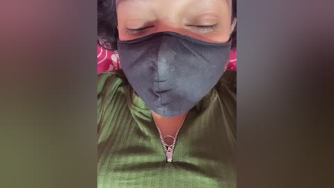 A video of a person with short, dark hair, wearing a dark face mask and green shirt, with a red and white patterned background, blurred.