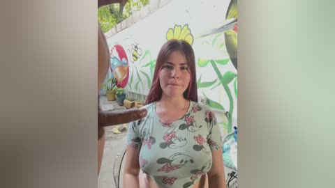 Video of a young woman with medium-length, dark brown hair, wearing a floral-patterned shirt, standing in a graffiti-covered alley. A man's hand is visible on the left, and the background shows colorful street art.