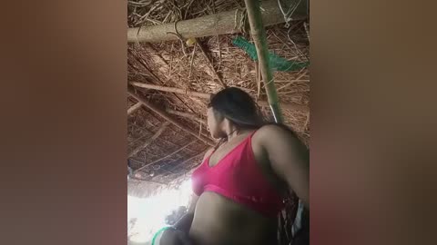 A video of a young woman with medium-dark skin and long black hair wearing a red tank top, standing inside a rustic hut with a thatched roof. The background features wooden beams and a dim, natural light filtering through.