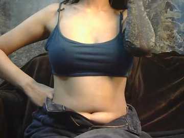 Video of a woman with medium skin tone, wearing a dark blue sports bra and unbuttoned jeans, sitting on a dark brown velvet couch, with a textured wall in the background.