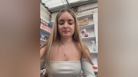 Video of a young woman with long, straight blonde hair, wearing a white off-the-shoulder top, standing in a cluttered, white-walled room with shelves and a sign reading \"Tobacco.\