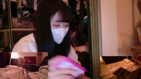Video of an Asian woman with long black hair and a white mask, seen through a mirror, in a dimly lit bedroom with clothes hanging and a messy bed.