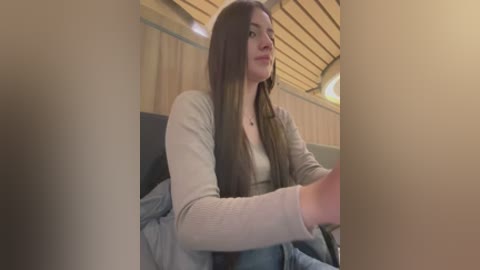 Video of a young Caucasian woman with long brown hair, wearing a beige sweater, seated in a cozy cabin with wooden walls and a soft, dim light.