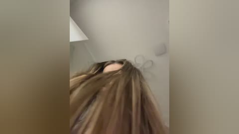 A blurred video of a woman with long, light brown hair, partially covering her face, standing in a dimly lit, beige-walled room with a ceiling lamp in the background.