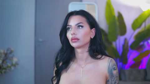 Video of a fair-skinned woman with long black hair, wearing a gold cross necklace, standing topless, revealing tattoos, in an indoor setting with a potted plant and blurred background.