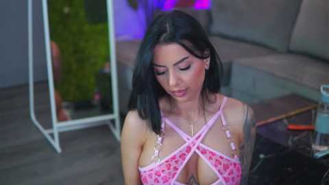 Video of a young woman with long black hair and light skin, wearing a pink, floral-patterned crop top with crisscross straps. She has medium-sized breasts. Background shows a modern indoor setting with plants and a glass table.
