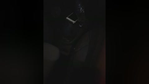 A dimly lit video of a person holding a camera, partially obscured by darkness, with a small light reflecting off the lens, creating a moody, slightly grainy atmosphere.