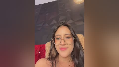 Video of a smiling Latina woman with long black hair and glasses, wearing a red top, against a geometric black and white wall, sitting on a textured fabric.