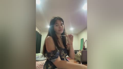 Video of a young Asian woman with long black hair, wearing a floral off-shoulder top, holding a glass, in a dimly lit bedroom with beige walls and a messy bed.