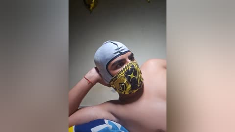 Video of a light-skinned person, likely male, with a grey headband, yellow and black face mask, and a blue and yellow shirt, lying on a bed with a white and blue blanket.